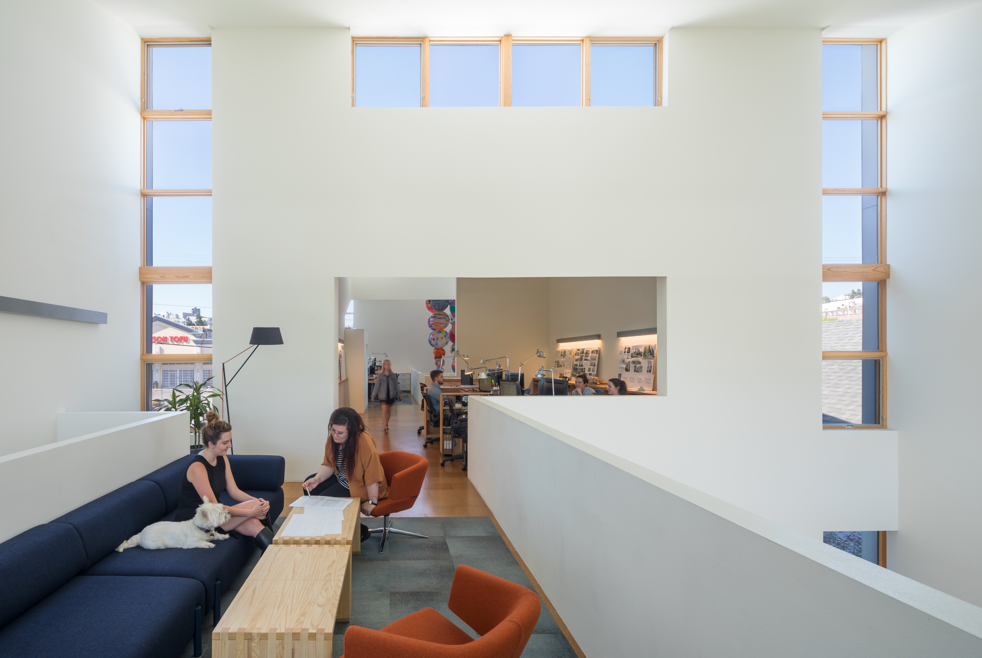 Office with blue couch and two orange chairs. Open concept meeting space shows desks with people working. The office has high ceilings and lots of windows with natural light.