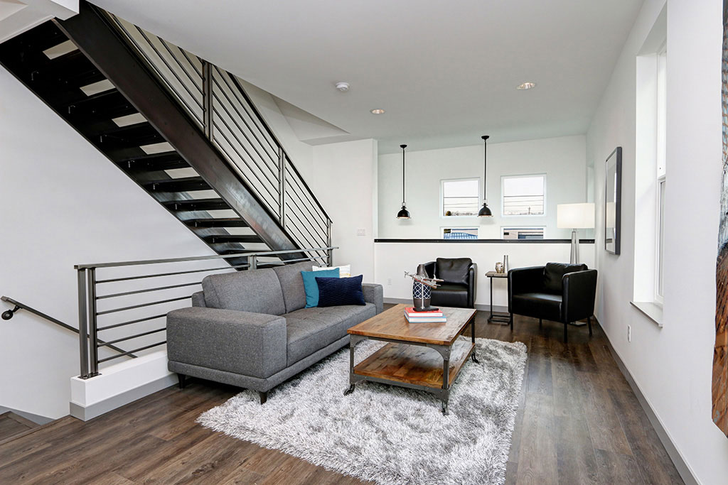Living room with dark wood floors and dark stairs, a gray couch, fuzzy rug, dark wood table, and two black leather chairs.