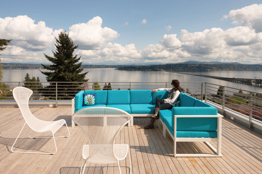 Rooftop deck with light wood flooring. Person sitting on bright turquoise outdoor couch, looking out at views of trees and water.