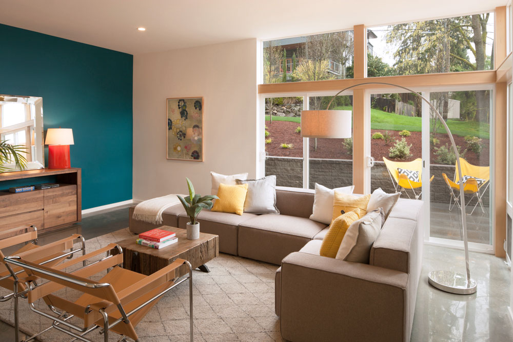 Living room with a beige couch with white and yellow pillows, two leather strap chairs, a wooden table, and a wooden credenza. One wall is dark teal and one wall is full of windows.