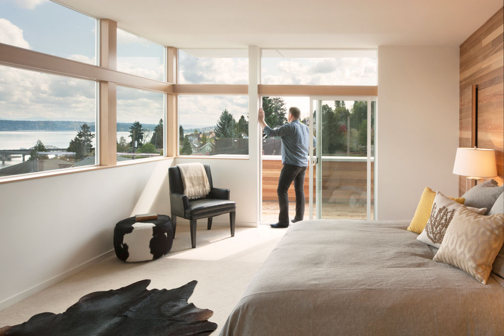 Man in bedroom, stepping outside through sliding doors to bedroom balcony. Bedroom is filled with natural light. Bedroom has a beige bed, an animal skin rug, and a cow print ottoman.
