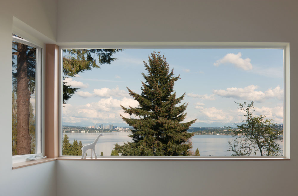 Large window with a large evergreen tree in the center of the window frame. Small porcelain white giraffe on window sill.