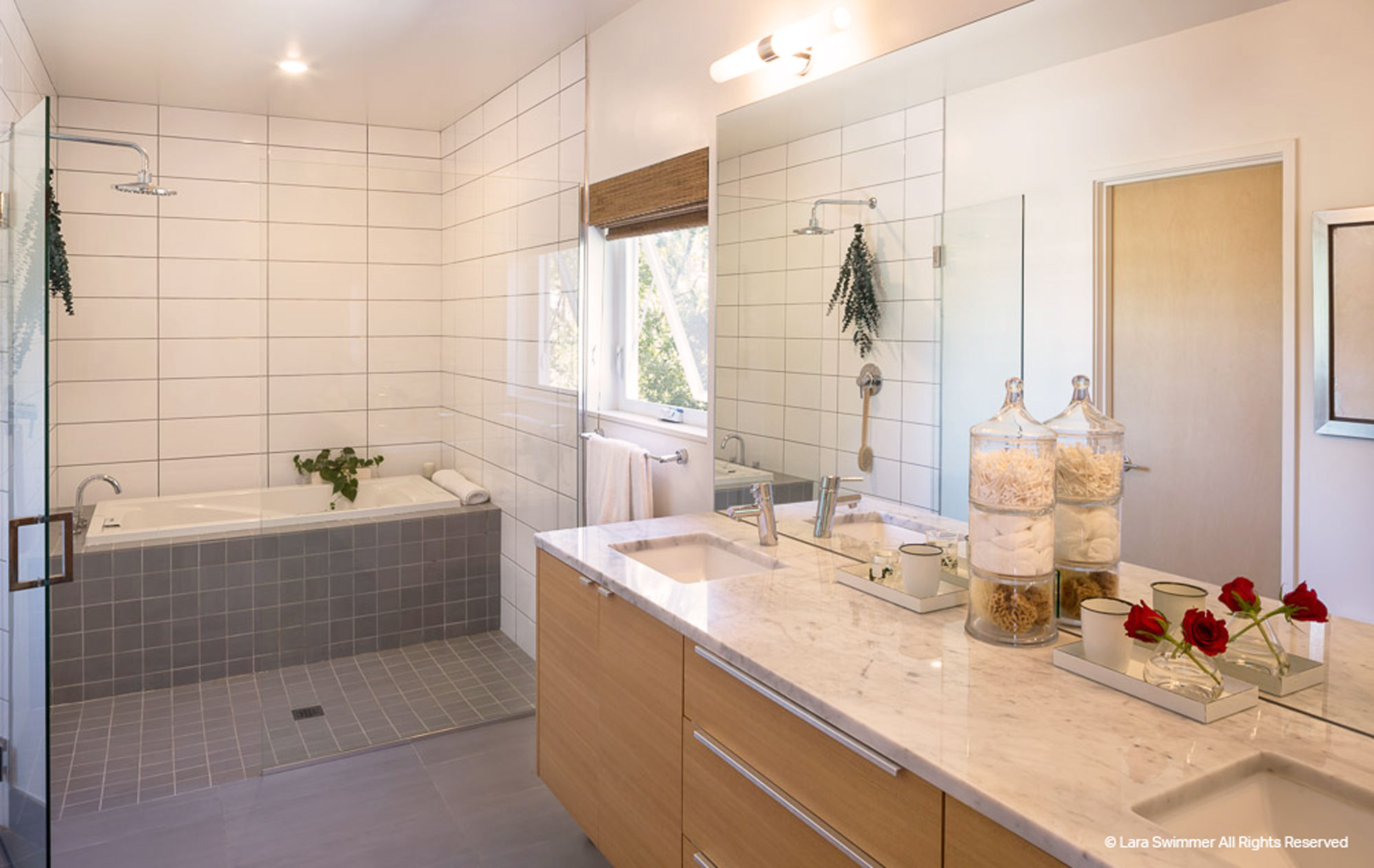 Bathroom with light wood cabinets and light marble counter tops. Gray tiles line bathtub.