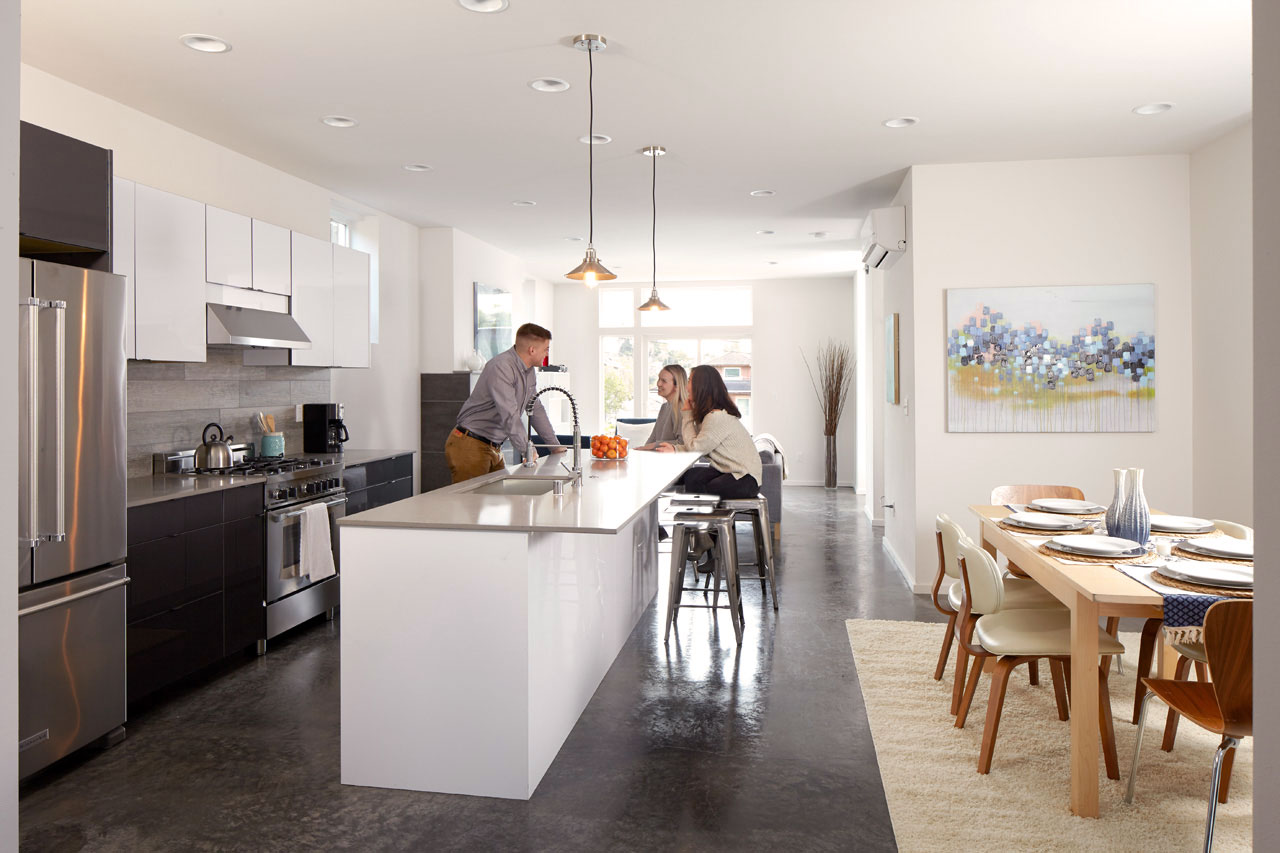 3 people around a white kitchen island with a silver top. Kitchen has a light wood table and chairs, a large painting with blue flowers, and silver kitchen fixtures.