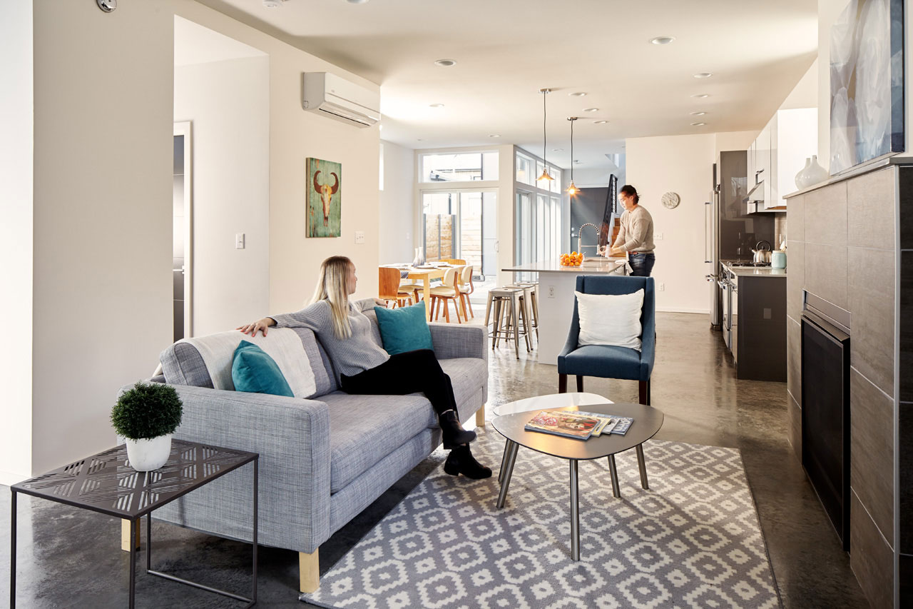 Interior photo of both the kitchen and living room. One person is sitting on a gray couch in front of a fireplace and one is visible and in the kitchen using the sink.