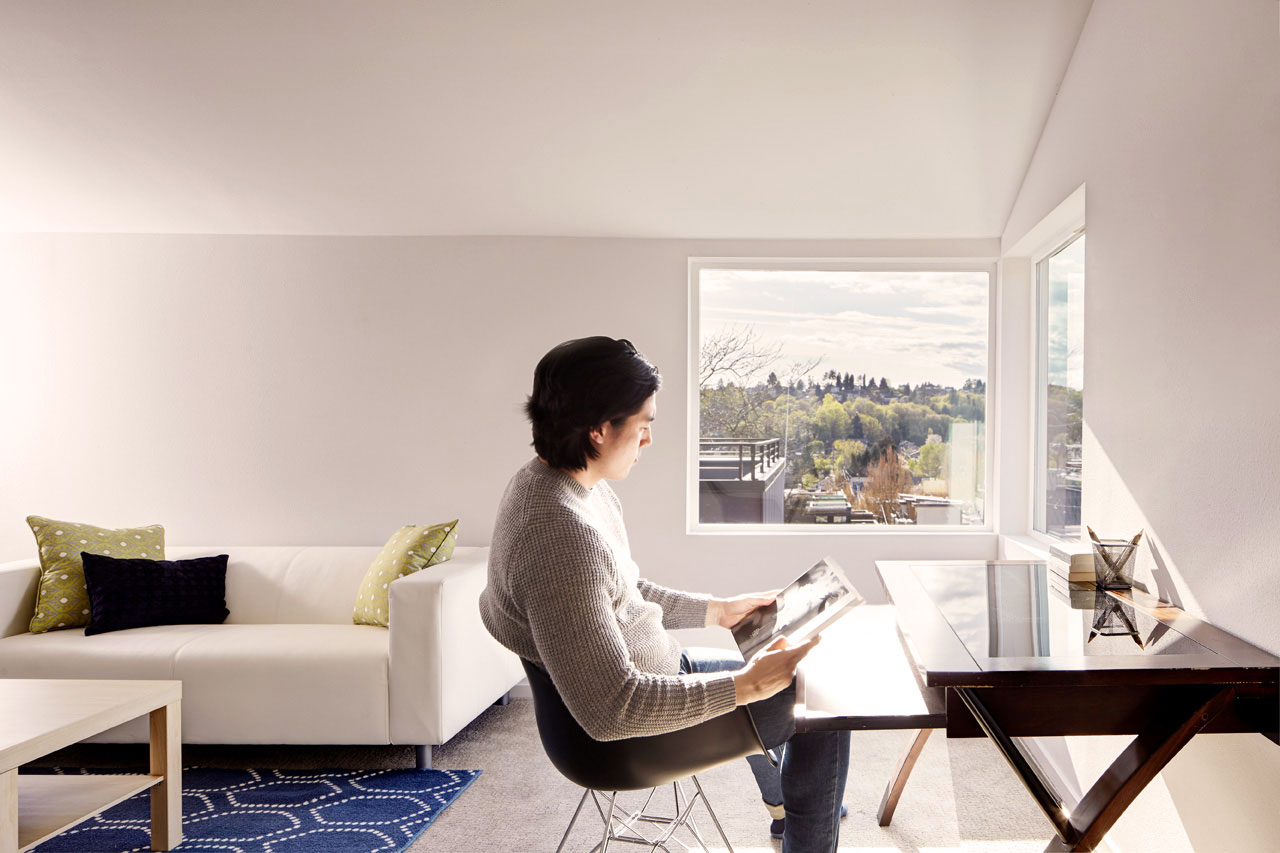 Person sitting at black desk reading in a living room with a white couch with lots of natural light coming from two windows