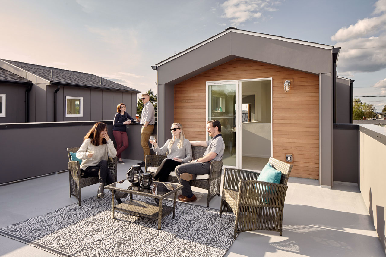 5 people on a rooftop deck, 3 are seated around an outdoor table, and two are in a corner by the door to the inside.