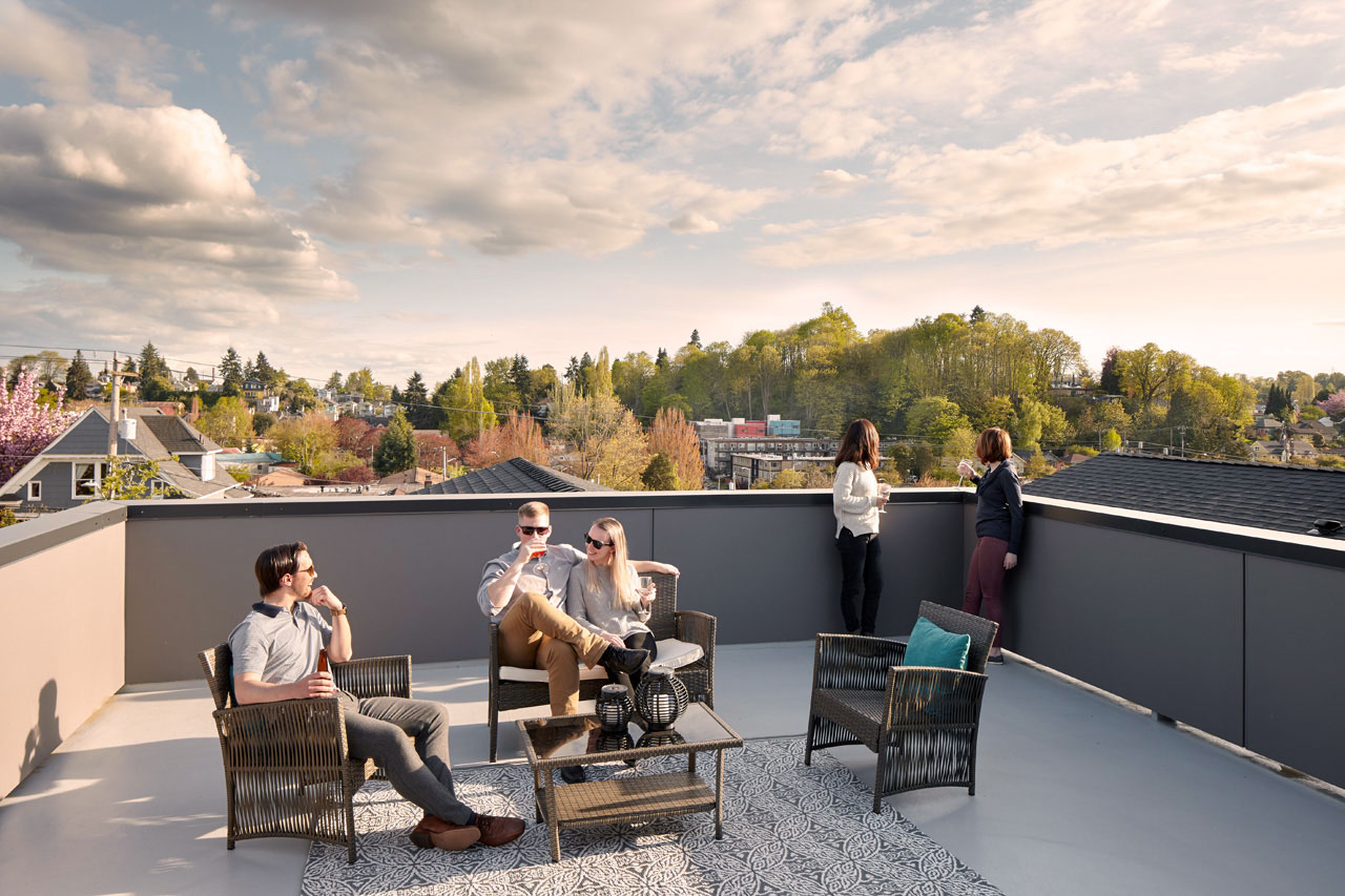 5 people on a rooftop deck, 3 are seated around an outdoor table, and two are looking off of the balcony at trees.