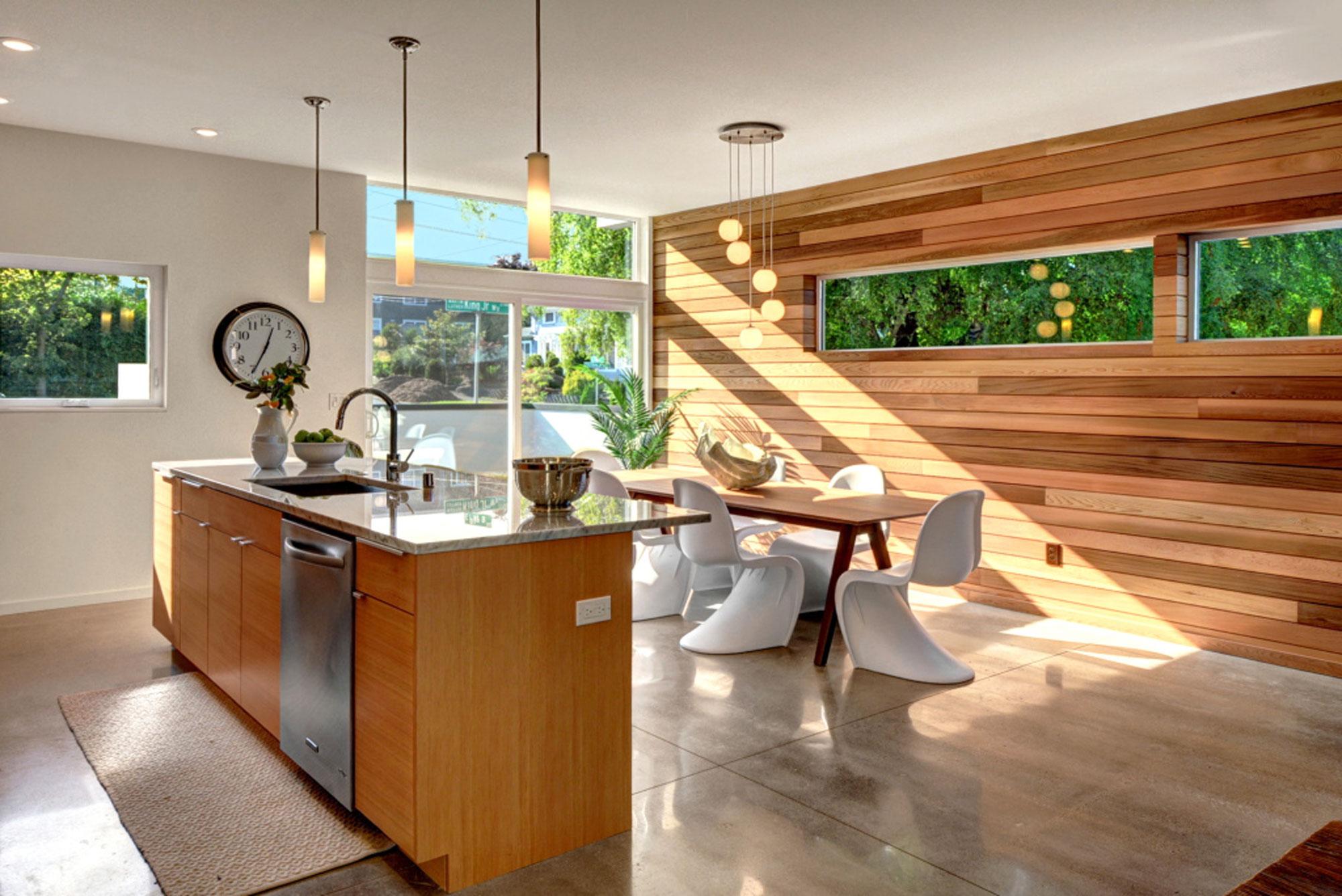 Kitchen with natural light pouring in from large floor to ceiling windows. Kitchen island is light wood with gray marble countertop. One wall of the kitchen is natural wood and the rest of the walls are white. There is a medium wood kitchen table with white mid century modern chairs.