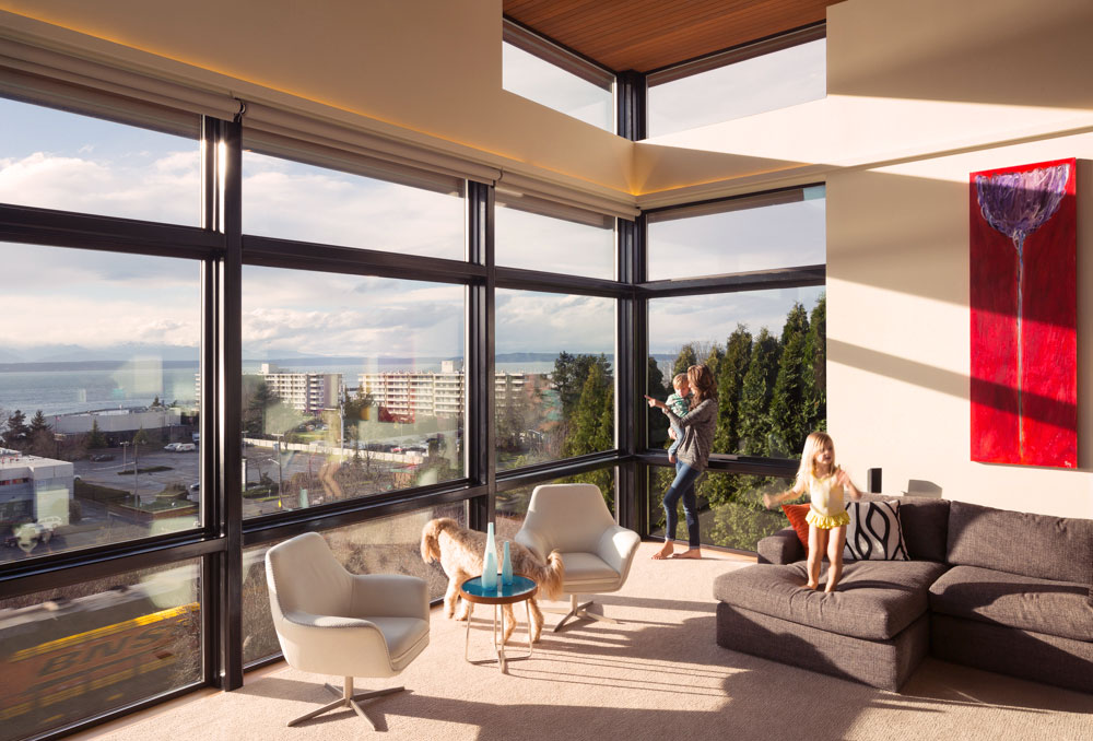 Living room with floor to ceiling windows. One person is holding a small child and looking out the one window, another child is standing on a gray couch. Golden doodle dog is also looking out of the window.