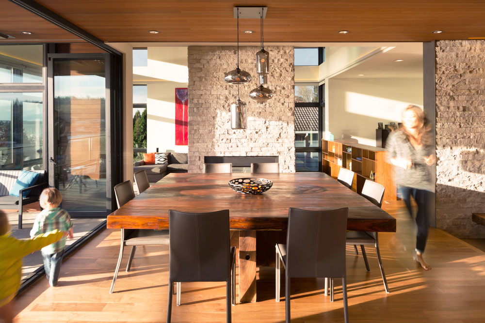 Dining room table with one adult and two children walking around the table. Open concept dining room with stone pillars between dining room and living room.
