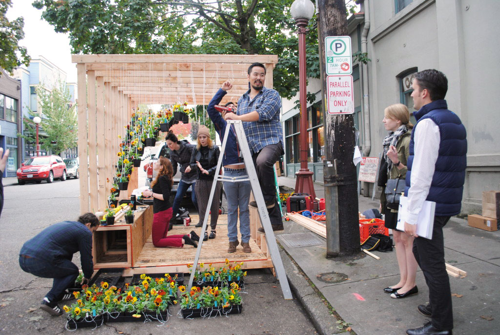 Group of people working together to string potted plants onto wooden structure. One person is on a ladder, others are behind stringing up plants