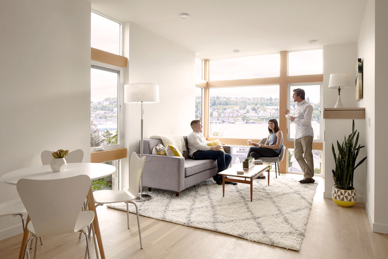 Mid century modern styled living room with one person sitting on a gray couch, one person sitting in a chair, and one person leaned up against a window. A kitchen table is also in view from the living room.